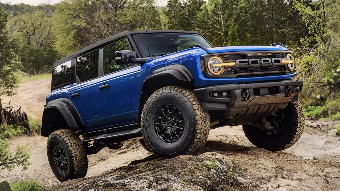 A blue Ford Bronco with a black roof and muddy wheels driving over rocky terrain.