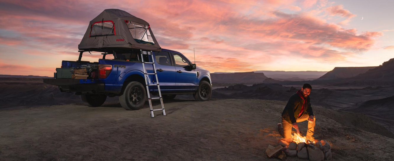A man camping in a desert by a campfire with a blue, Ford F-150 with a tent on top of it.