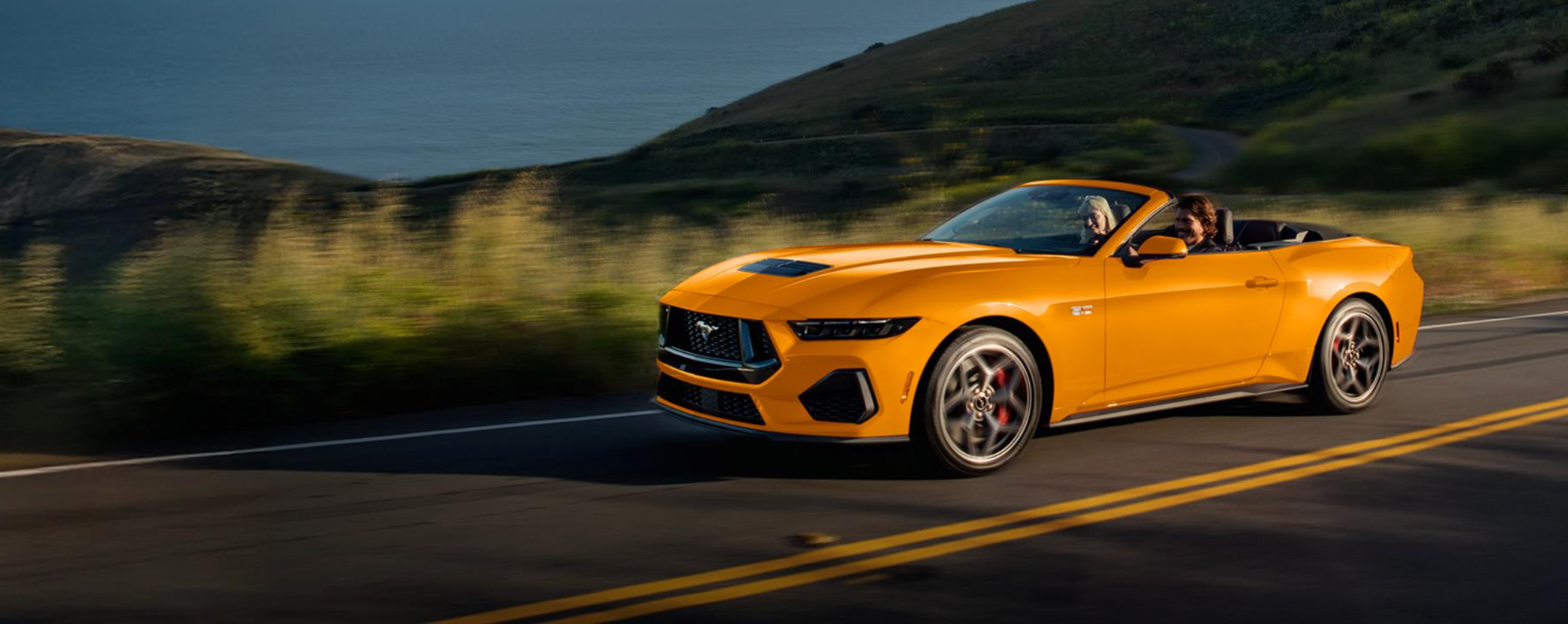 A yellow-orange Ford Mustang driving down the road in a hilly region.